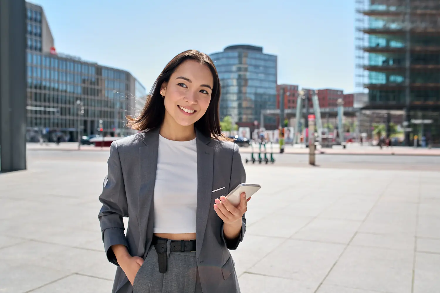 Young smiling elegant Asian busy business woman leader wearing suit standing in big city using cell phone platform applications. Smiling woman holding smartphone walking on street outdoors.