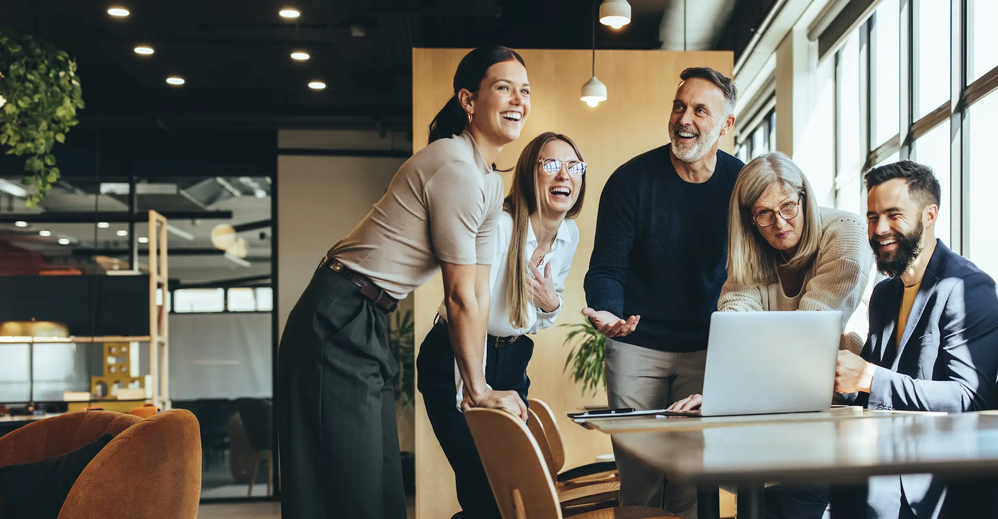 Happy businesspeople laughing while collaborating on a new project in an office. Group of diverse businesspeople using a laptop while working together in a modern workspace.